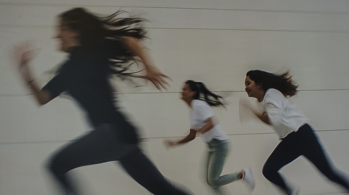 Three girls run in front of a white wall, their figures blurry with movement.
